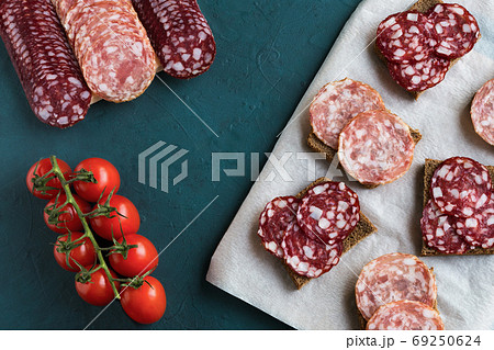Sandwiches with smoked traditional salchichon and braunschweig sausages are on board. Next to are several rows of sausages and cherry tomatoes on a green background. Flat lay, horizontal composition. Sandwiches with smoked traditional salchichon and braunschweig sausages are on board. Next to are several rows of sausages and cherry tomatoes on a green background. Flat lay, horizontal composition. 69250624