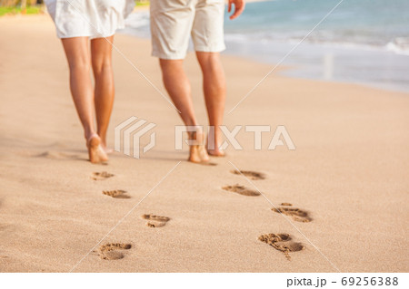Beach couple relaxing at sunset walking barefoot. Focus on footprints in golden sand. Closeup of legs. Romantic beach vacation holidays. Young people from behind walking away towards happiness. 69256388