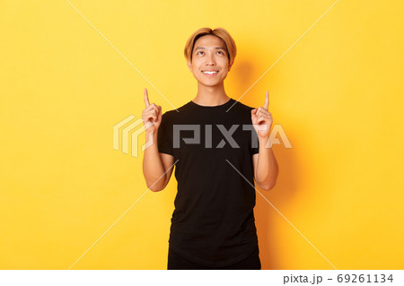 Handsome smiling asian man in black t-shirt pointing fingers up, looking at banner pleased, yellow background Handsome smiling asian man in black t-shirt pointing fingers up, looking at banner pleased, yellow background 69261134