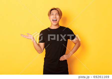 Portrait of happy blond asian guy, winking sassy and smiling, holding something on hand over yellow background 69261169