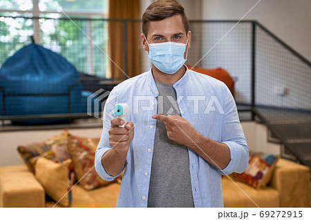 Working safely, portrait of young man wearing medical protective mask standing in the office and pointing at the infrared thermometer and looking at camera Working safely, portrait of young man wearing medical protective mask standing in the office and pointing at the infrared thermometer and looking at camera 69272915