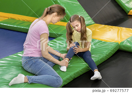 Trampoline jumping accident. Mom and child with foot injury sitting on rebounder at kids playground, indoor 69277577