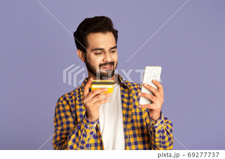 Cheerful Indian guy shopping online with mobile phone and credit card on violet background 69277737