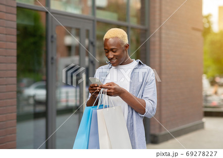 Smiling african american guy with packages with purchases in hands, typing on smartphone 69278227