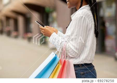 Meeting near store for shopping. Smiling african american woman blogger typing on smartphone and holding bags 69278250