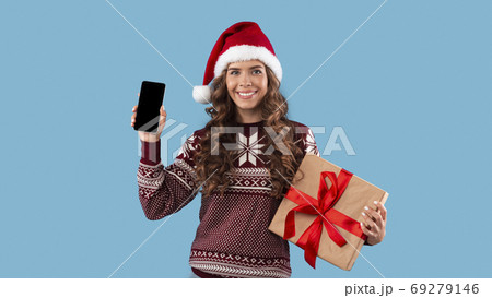Smiling girl in Christmas outfit holding gift box and cellphone with blank screen over blue background, space for design Smiling girl in Christmas outfit holding gift box and cellphone with blank screen over blue background, space for design 69279146