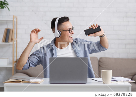 Guy in headphones sings at smartphone, sitting at table with laptop Guy in headphones sings at smartphone, sitting at table with laptop 69279229