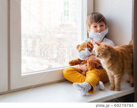 Toddler boy sits with teddy bear, both in medical masks. Kid with cute ginger cat. Fluffy pet and child on home quarantine because of coronavirus COVID-19. Toddler boy sits with teddy bear, both in medical masks. Kid with cute ginger cat. Fluffy pet and child on home quarantine because of coronavirus COVID-19. 69279783