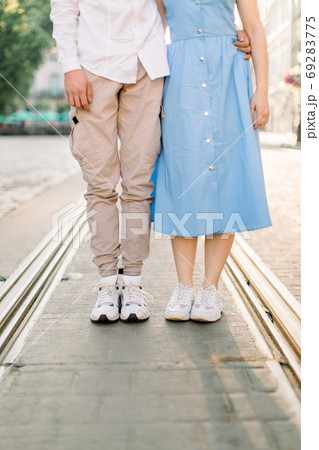 Cropped image of legs of lovely stylish young couple in love, woman in blue dress and man in beige pants, standing on the pavement road and tram track, having fun outdoors in the city 69283775