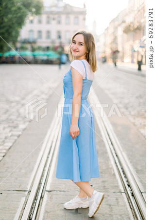 Portrait of fashionable beautiful Caucasian girl wearing blue dress and white sneakers, walking in old European city in a summer sunny day 69283791