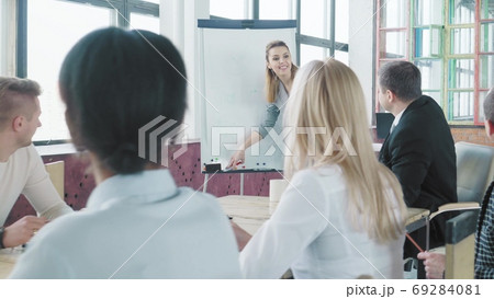 Business startup team holds a meeting at a large table. Girl manager gets up from a chair and walks over to a flip chart to start a presentation. Coworking. Office life Business startup team holds a meeting at a large table. Girl manager gets up from a chair and walks over to a flip chart to start a presentation. Coworking. Office life 69284081
