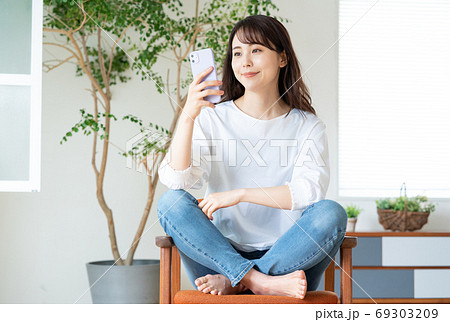 リビングで椅子に座ってスマホを操作する笑顔の若い女性 69303209