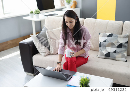 Woman is sitting on the couch holding cup in crayfish and with her other hand typing on laptop keyboard. 69311222