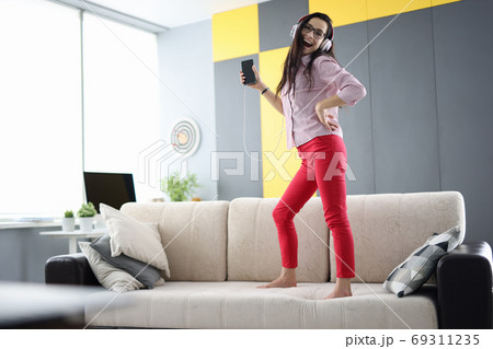 Woman is standing on couch with headphones laughing and holding smartphone in her hands Woman is standing on couch with headphones laughing and holding smartphone in her hands 69311235