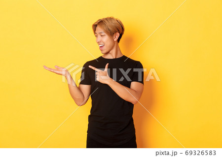Portrait of handsome smiling asian guy holding something on hand and pointing finger at it, standing yellow background pleased Portrait of handsome smiling asian guy holding something on hand and pointing finger at it, standing yellow background pleased 69326583