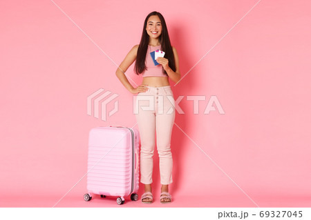 Travelling, holidays and vacation concept. Full-length of cheerful smiling asian girl tourist with suitcase, holding passport with flight tickets, heading to aiport to start journey, pink background Travelling, holidays and vacation concept. Full-length of cheerful smiling asian girl tourist with suitcase, holding passport with flight tickets, heading to aiport to start journey, pink background 69327045