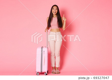 Travelling, holidays and vacation concept. Full-length of coquettish dreamy asian girl tourist enjoying tour, pointing finger and looking up enthusiastic, standing with suitcase over pink wall 69327047