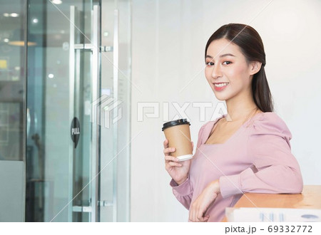 Young beautiful asian woman in pink  shirt holding paper cups hot beverage or coffee,standing and smiling in the office before starting work in the morning 69332772