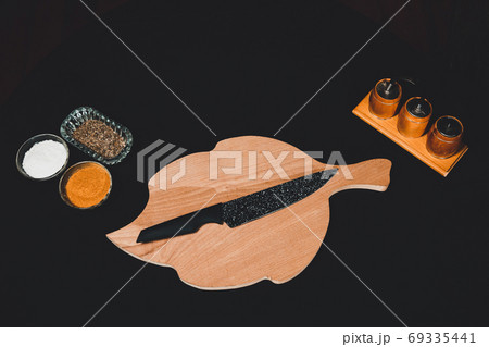 On a black background a wooden board with a black knife and a set of spices on kitchen utensils. 69335441