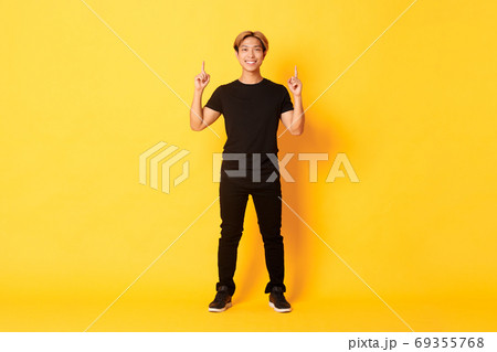 Full-length of confident handsome asian man with blond hair, smiling and pointing fingers up, showing logo, yellow background Full-length of confident handsome asian man with blond hair, smiling and pointing fingers up, showing logo, yellow background 69355768