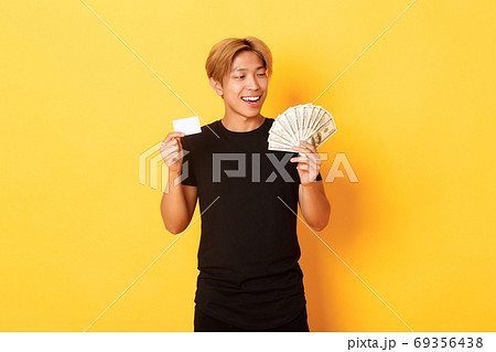 Happy and lucky asian guy looking pleased at money while showing credit card and cash, standing yellow background Happy and lucky asian guy looking pleased at money while showing credit card and cash, standing yellow background 69356438