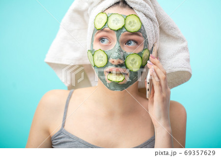 Woman with towel on head and face mask enriching with cucumbers via smartphone Woman with towel on head and face mask enriching with cucumbers via smartphone 69357629