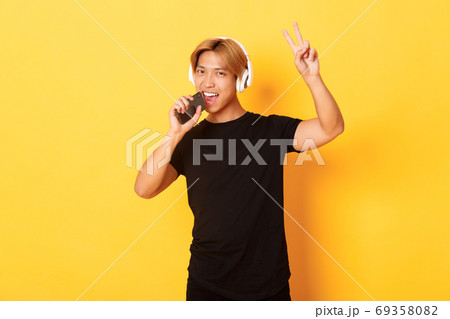 Sassy handsome asian guy in headphones, playing karaoke app, singing into mobile phone microphone, showing peace gesture, standing yellow background Sassy handsome asian guy in headphones, playing karaoke app, singing into mobile phone microphone, showing peace gesture, standing yellow background 69358082