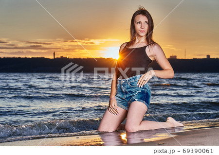 A young brunette girl in a black T-shirt and denim shorts kneels on the sandy shore A young brunette girl in a black T-shirt and denim shorts kneels on the sandy shore 69390061