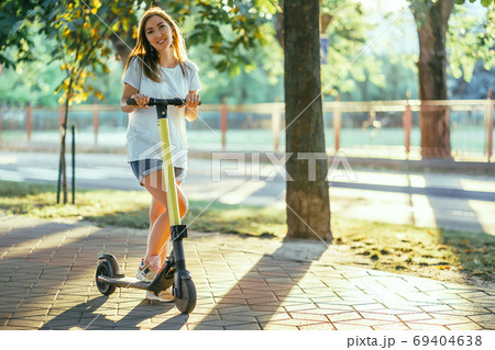 Smiling woman rides electric scooter or e-scooter in city park at sunset. Female using electric transport in urban park Smiling woman rides electric scooter or e-scooter in city park at sunset. Female using electric transport in urban park 69404638