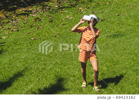 Young woman testing VR eyeglasses outside in tropical garden. Female in bright orange summer outfit wearing virtual reality headset during summer. Active modern futuristic leisure lifestyle Young woman testing VR eyeglasses outside in tropical garden. Female in bright orange summer outfit wearing virtual reality headset during summer. Active modern futuristic leisure lifestyle 69423162