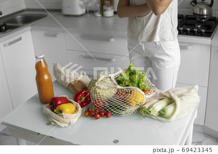 Woman came back from a market and unpacks a reusable grocery bag full of vegetables on a kitchen at home. Zero waste and plastic free concept. Girl is holding mesh cotton shopper with vegetables. Woman came back from a market and unpacks a reusable grocery bag full of vegetables on a kitchen at home. Zero waste and plastic free concept. Girl is holding mesh cotton shopper with vegetables. 69423345