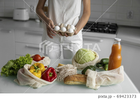 Woman holding eggs tray with a reusable shopping bags with vegetables on a table on a kitchen at home. Zero waste and plastic free concept. Mesh cotton shopper with vegetables. Ecology. 69423373