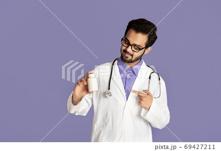 Confident Indian doctor pointing at jar of pills on lilac background, mockup for design 69427211