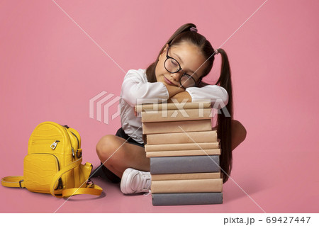 Sad and tired concept. Girl in glasses in uniform with backpack sleeps on stack of books Sad and tired concept. Girl in glasses in uniform with backpack sleeps on stack of books 69427447