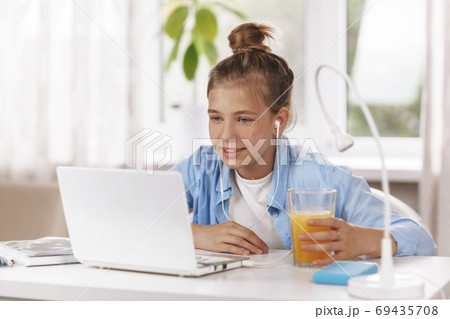 Teenage girl wearing headphones sitting at table and doing homework by video conference using laptop Teenage girl wearing headphones sitting at table and doing homework by video conference using laptop 69435708