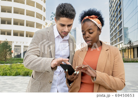 Young elegant businessman magnifying photo in smartphone of African colleague Young elegant businessman magnifying photo in smartphone of African colleague 69450653