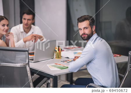 Bearded male sitting at table opposite his male and female coworkers Bearded male sitting at table opposite his male and female coworkers 69450672