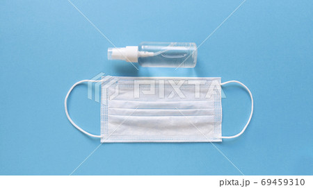 White medical mask and hand sanitizer in transparent bottle with spray cap at the middle of blue background. Simple flat lay with pastel paper texture. Medical concept. Stock photo. White medical mask and hand sanitizer in transparent bottle with spray cap at the middle of blue background. Simple flat lay with pastel paper texture. Medical concept. Stock photo. 69459310