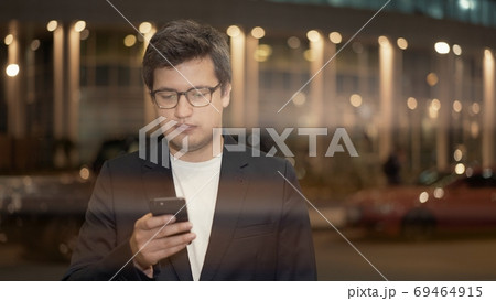 Businessman in eyeglasses scrolling the phone on background of building, lens flare 69464915