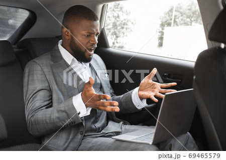 Angry black businessman with laptop going by taxi to job Angry black businessman with laptop going by taxi to job 69465579