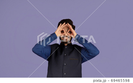 Portrait of comic Indian man holding fingers near his eyes like glasses, violet background 69465598