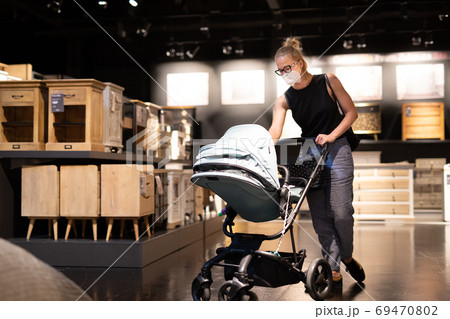 Young mom with newborn in stroller shopping at retail furniture and home accessories store wearing protective medical face mask to prevent spreading of corona virus. New normal during covid epidemic Young mom with newborn in stroller shopping at retail furniture and home accessories store wearing protective medical face mask to prevent spreading of corona virus. New normal during covid epidemic 69470802