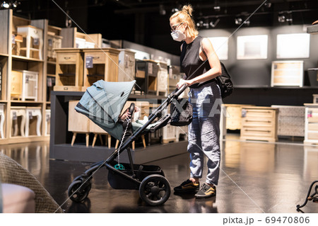 Young mom with newborn in stroller shopping at retail furniture and home accessories store wearing protective medical face mask to prevent spreading of corona virus. New normal during covid epidemic Young mom with newborn in stroller shopping at retail furniture and home accessories store wearing protective medical face mask to prevent spreading of corona virus. New normal during covid epidemic 69470806