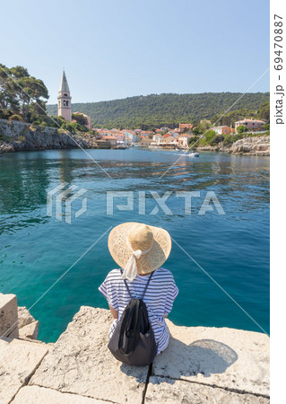 Woman traveler wearing straw summer hat and backpack, sittingat edge of stone pier, enjoying beautiful panoramic view of Veli Losinj, Losinj island, Croatia. Woman traveler wearing straw summer hat and backpack, sittingat edge of stone pier, enjoying beautiful panoramic view of Veli Losinj, Losinj island, Croatia. 69470887