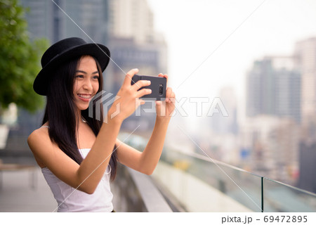 Happy young beautiful Asian tourist woman taking picture with phone against view of the city 69472895