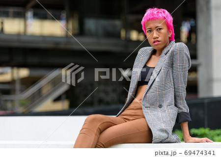Young rebellious Asian businesswoman with pink hair sitting in the city 69474341