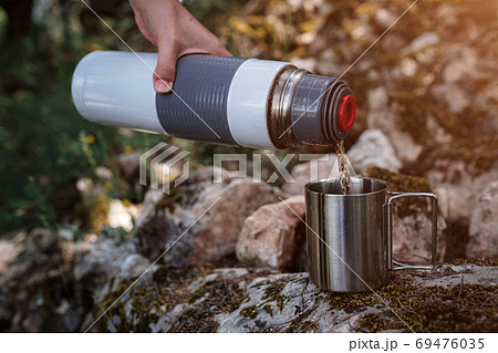 In a camping mug pours tea from a thermos in nature. In a camping mug pours tea from a thermos in nature. 69476035