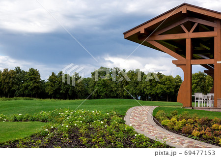 A hilly landscape with a flowerbed mulching for roses and footpath leading to a wooden canopy of a street cafe terrace in a park with a green forest in the background. 69477153