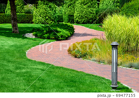 a winding pedestrian walkway made of red paving slabs with a gray stone curb in a park with a green lawn and reeds illuminated by sunlight, a ground street light was installed on the lawn. 69477155
