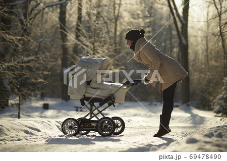 Young woman in coat raised on tiptoes, bending over baby stroller, looking at child. Sunny winter park, childcare, love 69478490